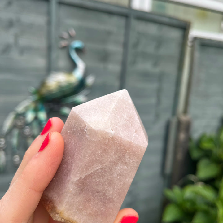 A close-up view of the Pink Amethyst crystal tower angled softly between fingertips with painted nails. The smooth, polished surfaces and heavy mineral textures are fully captured, making it a supportive companion to encourage emotional balance, grace, and inner peace.
