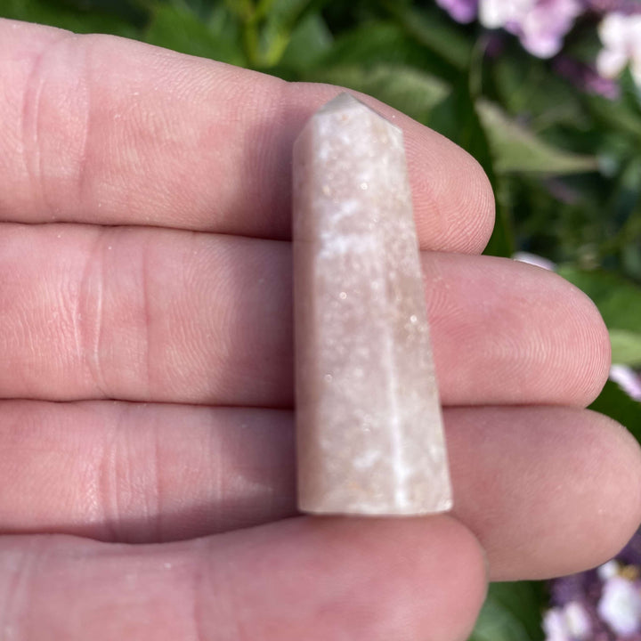 An overhead shot looking down at the pale pinkish-beige amethyst crystal tower resting flat against an open hand outdoors. The polished, slick facets catch the natural morning sun, emphasizing its organic, light-coloured patterns to help create a cosy sanctuary for daily relaxation.
