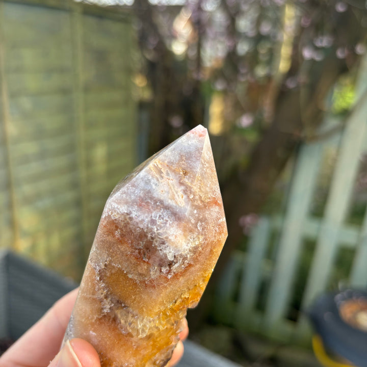 A hand tilting the pink amethyst crystal tower at a sharp angle outdoors, showing the smooth, light-reflective facets and intricate geological birthmarks, ideal for styling on a cosy bedside layout.