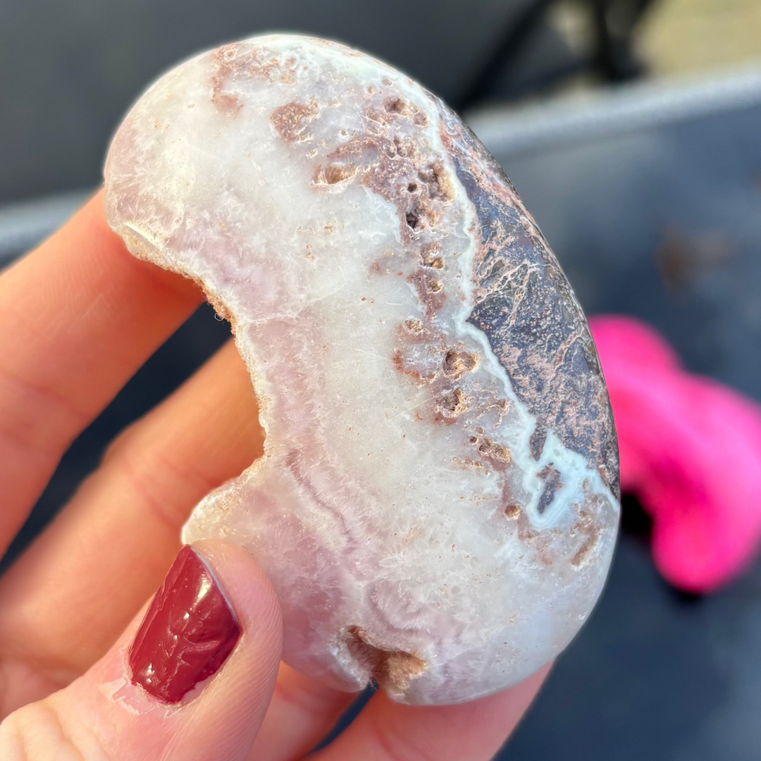 A close-up view of a unique, crescent-shaped Pink Aragonite palm stone held upright in a hand with painted fingernails. The polished healing crystal showcases intricate rose-cream layers and natural surface pits, beautifully highlighting its dual Root and Heart Chakra alignment.