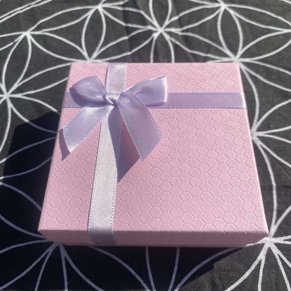 A high-angle shot of the closed, square light pink gift box highlighting its textured honeycomb exterior. The packaging is decorated with a neatly tied satin lilac ribbon and bow, presented flat against a contrasting black fabric background with a white geometric line pattern.