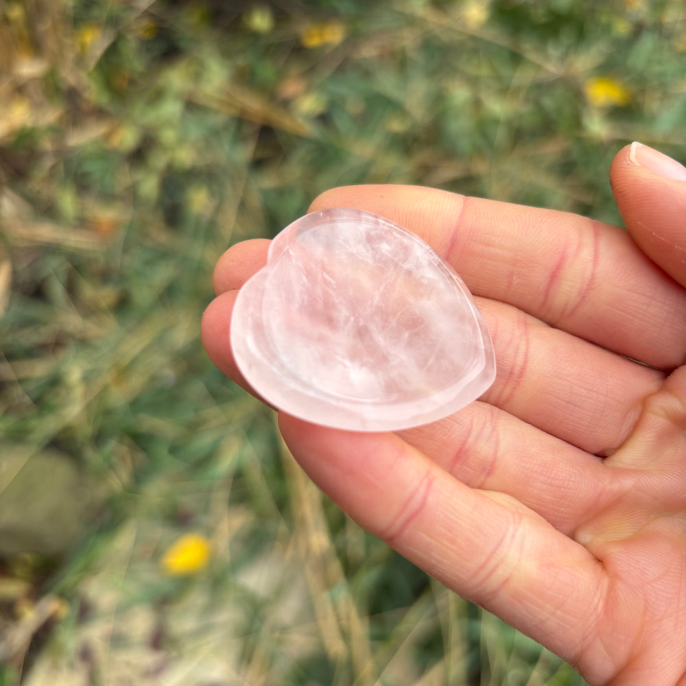 A hand holding a translucent pink quartz crystal thumb stone at an angle to reveal its central indented groove for anxiety relief.