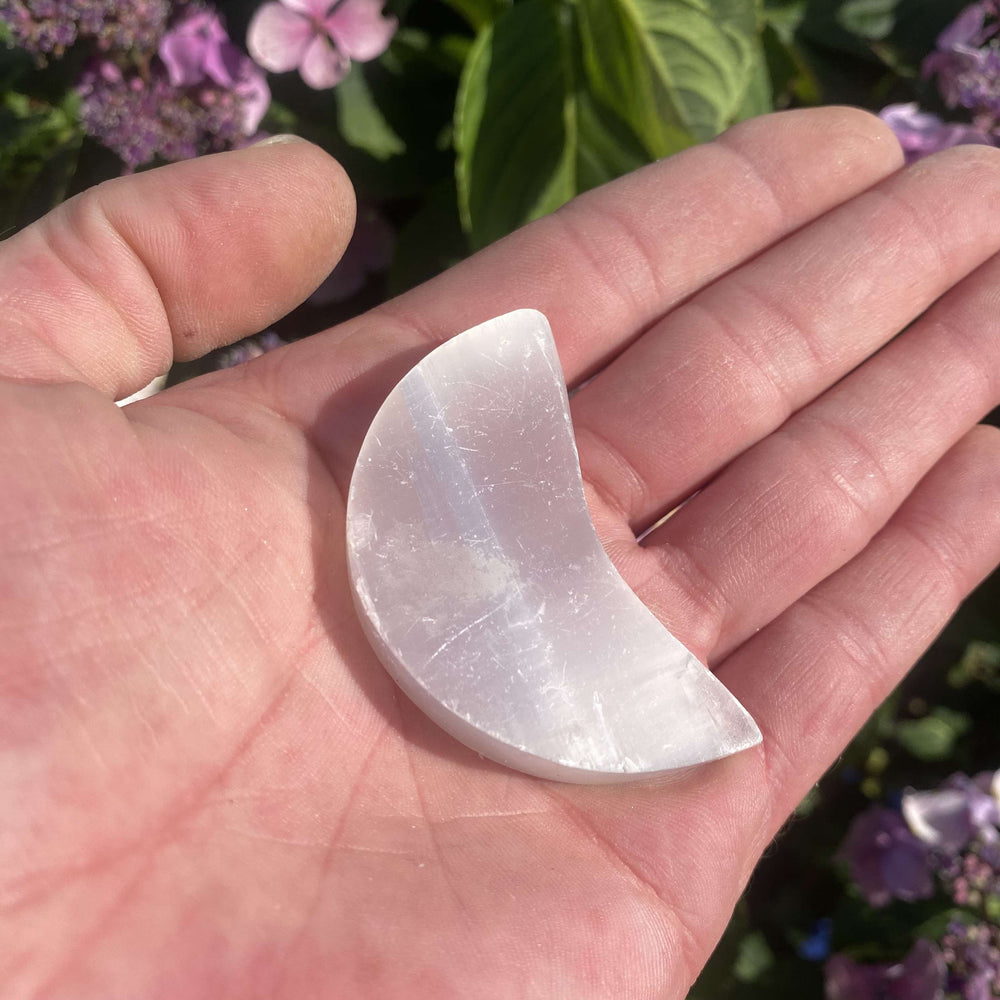 An outdoor shot of the smooth white Selenite crystal moon carving resting in an open hand from a distance, illustrating its pocket-sized palm scale and translucent appearance.