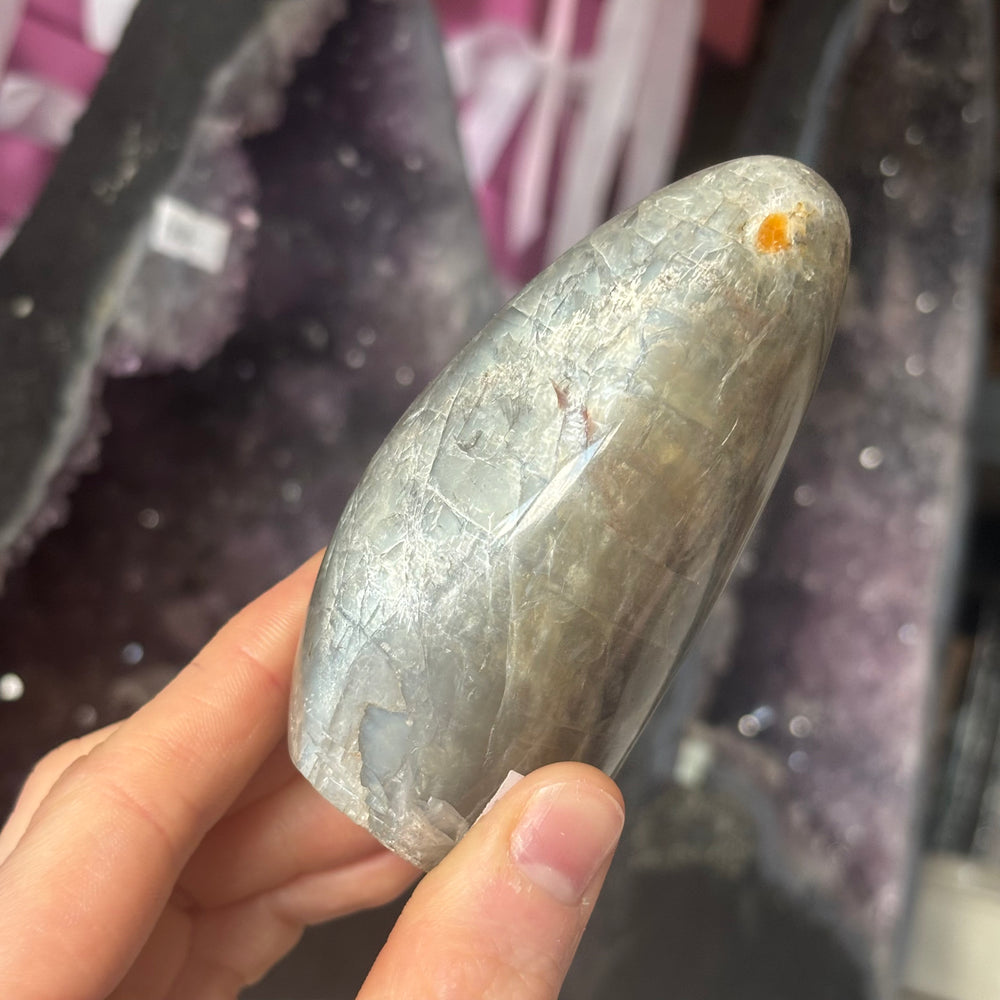A hand holding the self-standing Black Moonstone freeform crystal to demonstrate its solid weight and smooth texture. The background shows a large amethyst geode, highlighting how this piece serves as a magical companion for bedside tables or altars.