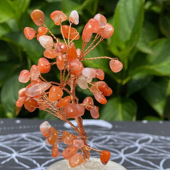 A detailed close‑up shot focusing on the delicate copper branches and vibrant, sunset‑hued Carnelian gemstone chips catching natural daylight against a blurred leaf background.