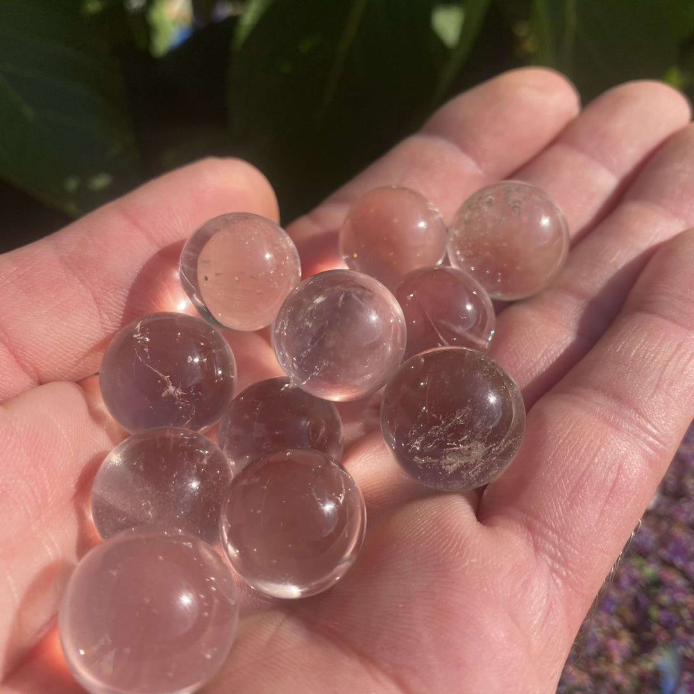 A pile of tiny, perfectly round Clear Quartz orbs cradled in an open palm outdoors under bright daylight. This angle highlights the natural glass-like reflections, subtle internal veils, and icy inclusions running through the genuine mineral gemstones.