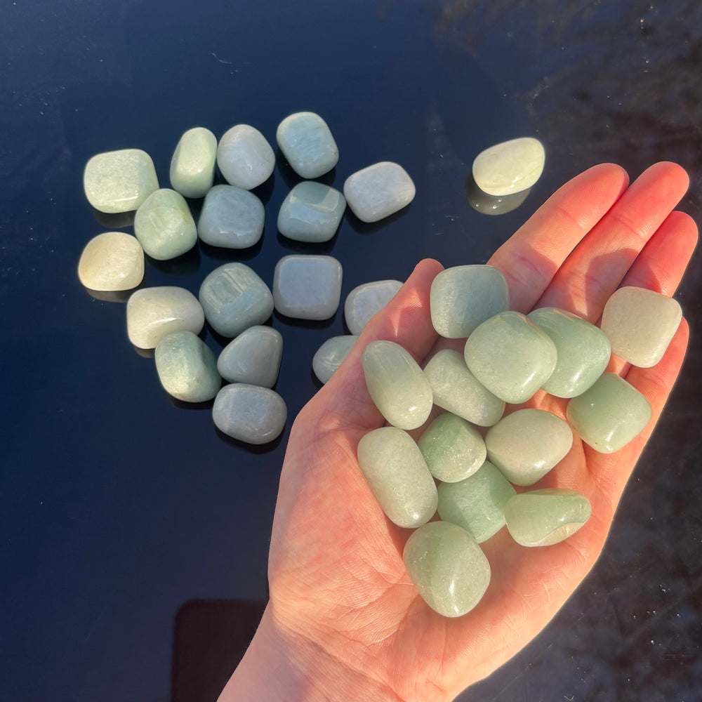 A hand holding a generous handful of polished green aventurine tumbles outdoors next to a pile scattered on a dark background layout, highlighting the unique natural markings and light variations of these good fortune stones.