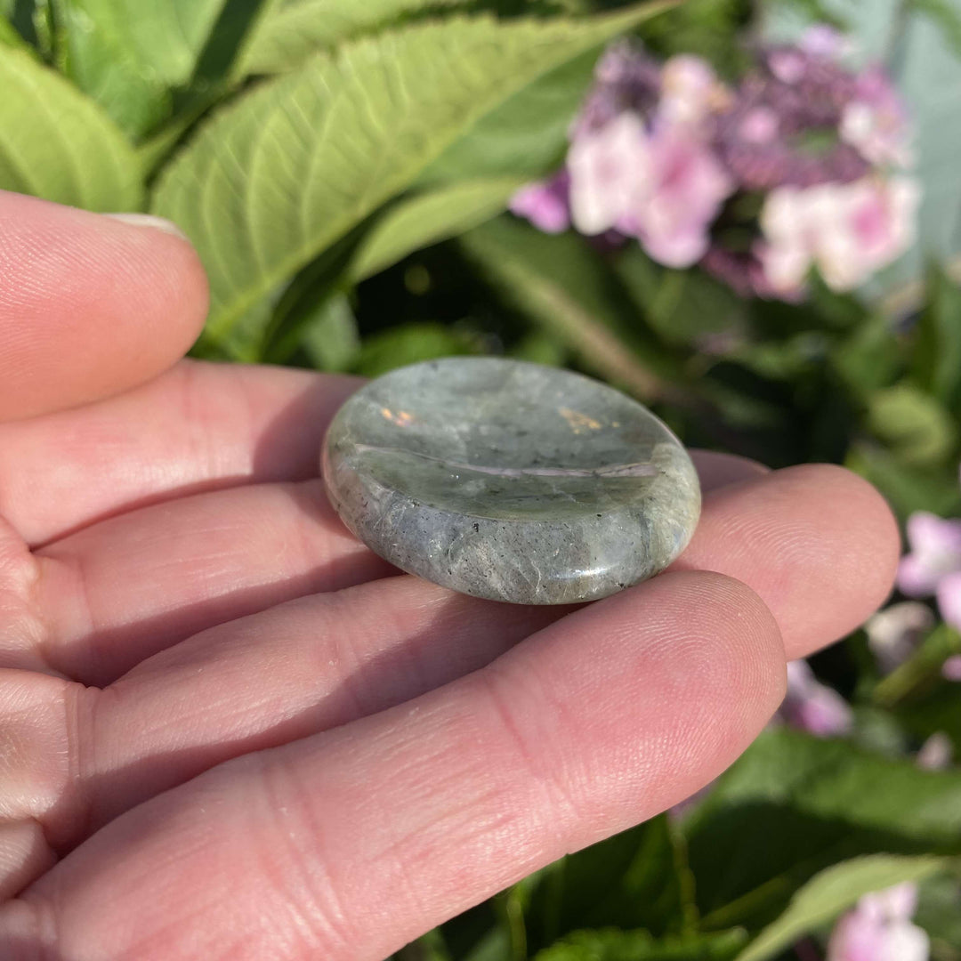 A close-up view looking down at a light-coloured gray Labradorite pocket friend resting flat on a palm outdoors. The natural morning sun accentuates its tactile, smooth finish and unique mineral landscapes, creating a cosy, supportive anchor for emotional balance and gentle personal transitions.