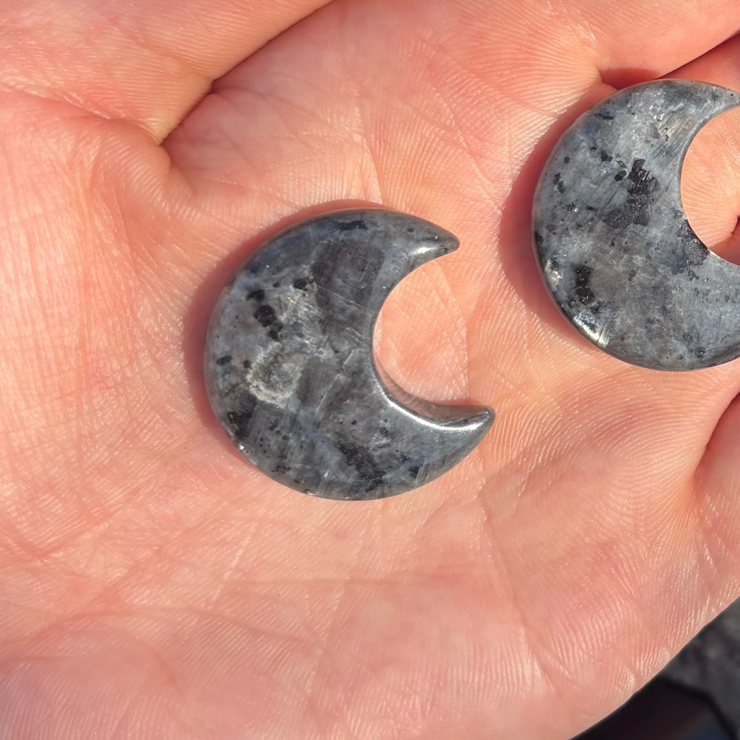 A close-up view focusing on two polished Larvikite crescent moon carvings resting flat on fingers under the morning sun. The sweeping curves and smooth edges of each celestial silhouette are fully visible, showcasing hidden silvery blue flashes that bring a quiet sense of wonder.