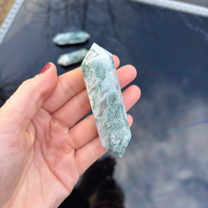 A close-up view of a green Moss Agate double terminated healing crystal held vertically to emphasize its smooth, pocket-sized therapeutic design.