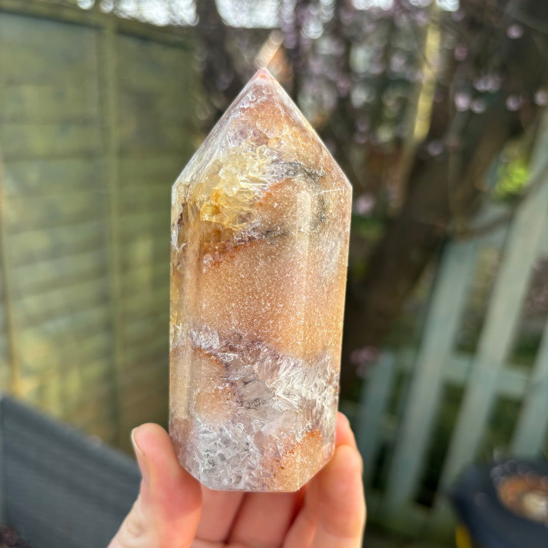 A hand displaying a rotated face of the polished pink amethyst obelisk tower outdoors, showcasing the beautiful dustings of natural blush colour tones that bring a soft reset to emotional stress.
