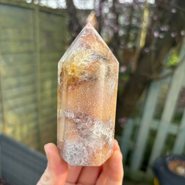 A hand displaying a rotated face of the polished pink amethyst obelisk tower outdoors, showcasing the beautiful dustings of natural blush colour tones that bring a soft reset to emotional stress.
