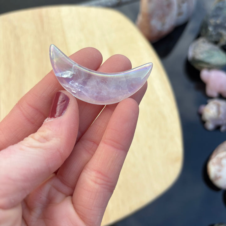 A close-up view of a single polished pink aura quartz moon carving held up against a dark outdoor setting, detailing its smooth curved edges and iridescent rainbow sheen that encourages deep emotional healing.