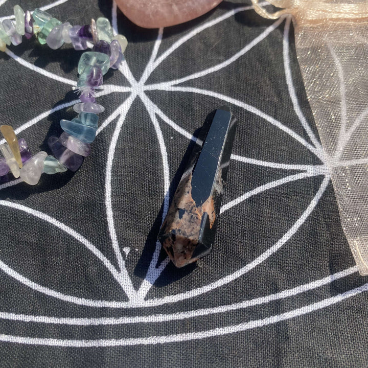 A macro shot focusing on the polished rhodonite crystal generator point beside a sparkling fluorite chip bracelet. The dark rosy tones of the tower point catch the sun next to the iridescent purple and green gemstone jewelry beads.