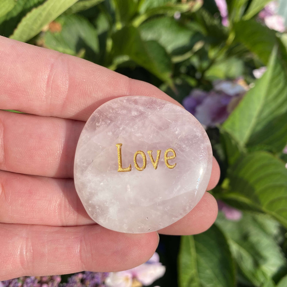 A close-up view of a heart-centred Rose Quartz palm stone cradled flat on a hand outdoors. The gold-engraved lettering catches the soft morning sun, showcasing its delicate inner layers and comforting pink hues that promote deep self-love, warmth, and emotional ease within a cosy space.