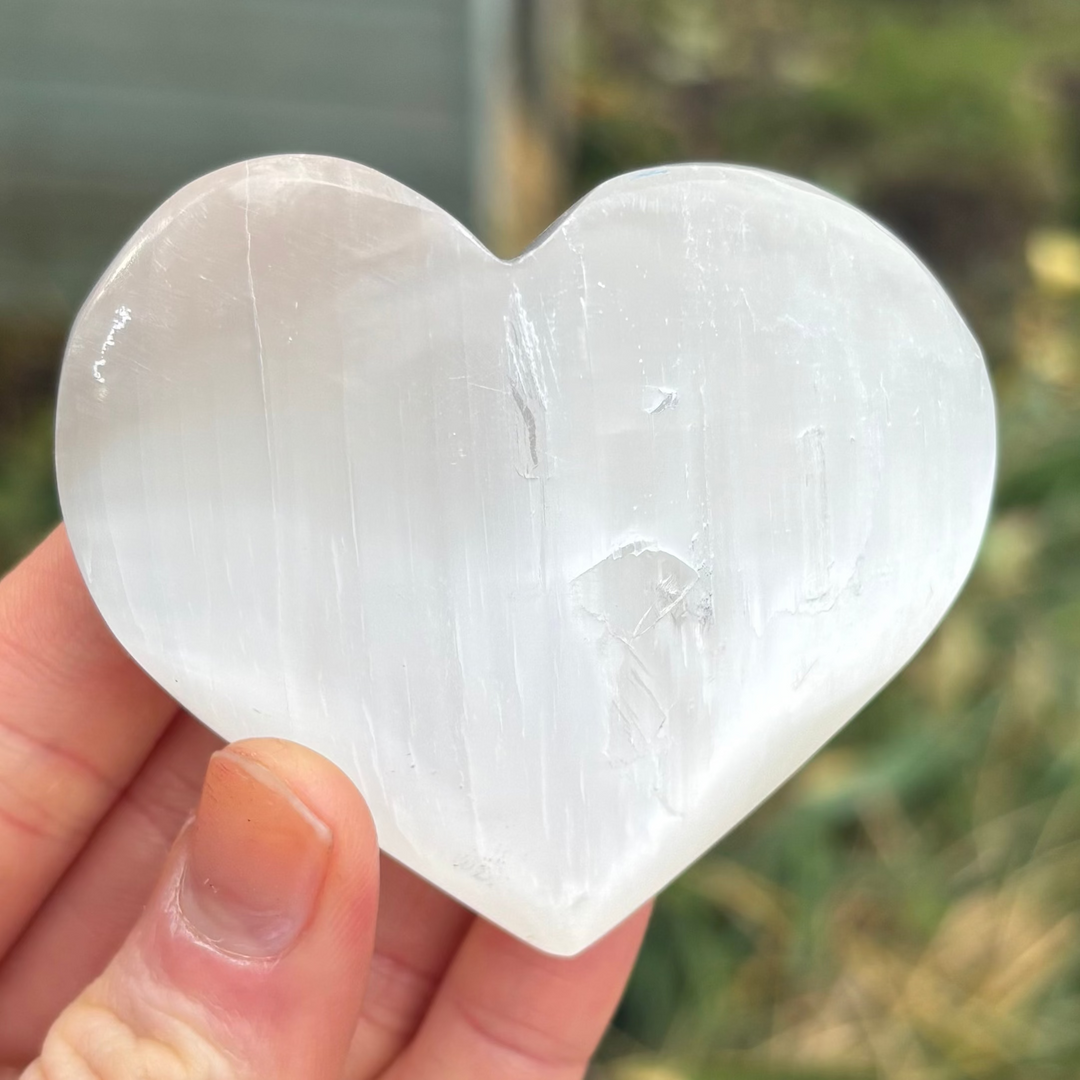 A close-up view of a single polished white selenite puff heart held between fingers, detailing the sweeping vertical grain fibers and radiant pearlescent flash that brings a deeply soothing aura to mindfulness practices.
