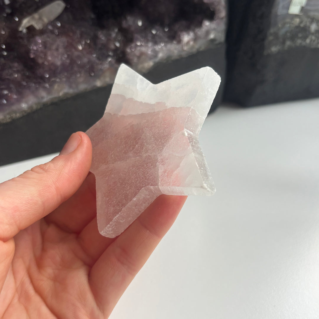 A close-up shot of fingers tilting the polished white selenite star carving sideways to showcase its smooth cut edge profile, flat thickness, and natural inner frost lines designed to clear away stagnant vibes.