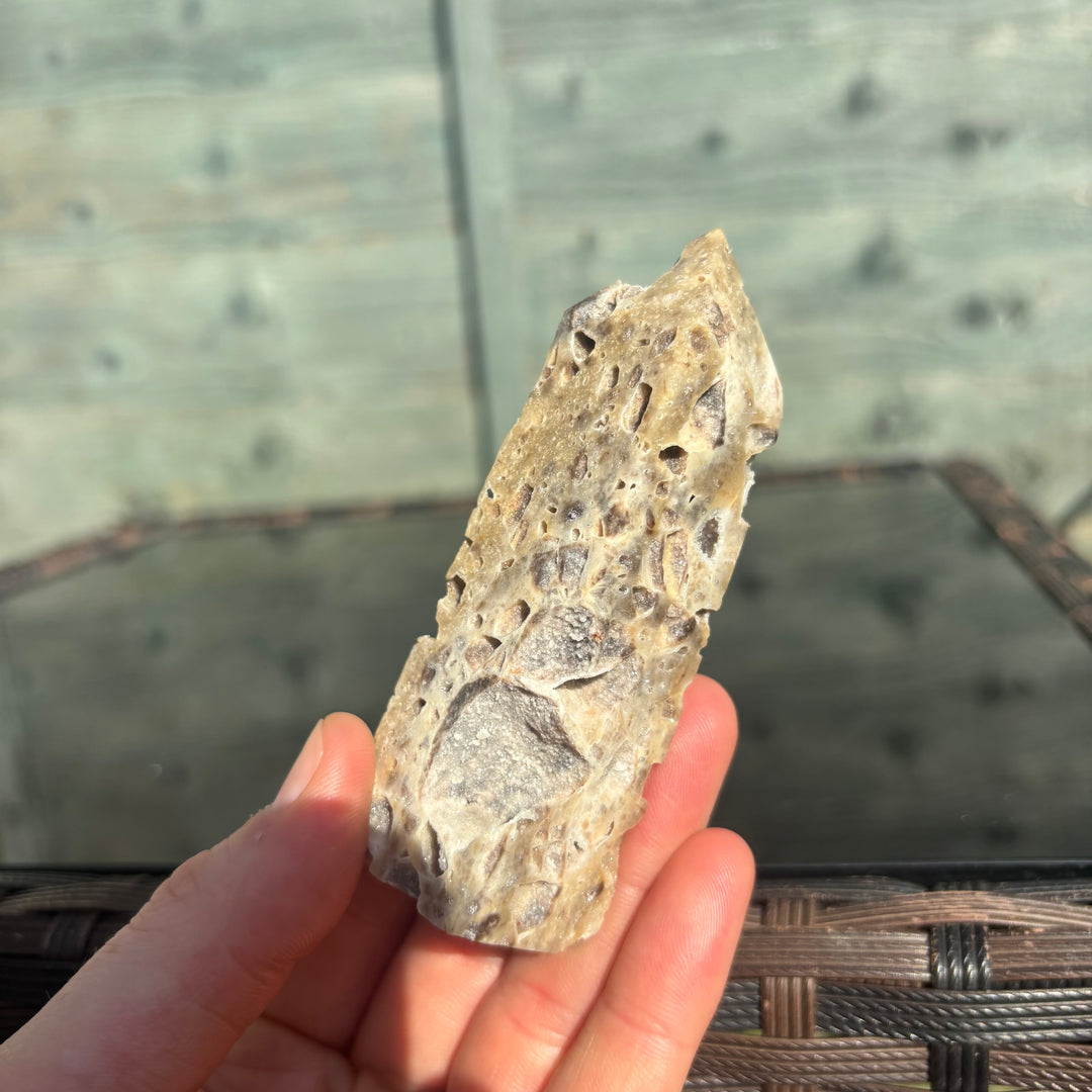 A close-up view looking down at a hand lifting the heavy Sphalerite crystal tower from its display surface to showcase its true real-world scale. The light-coloured, shimmering patterns and organic textures are fully visible, offering a supportive companion designed to unlock fresh creativity and align with the Sacral Chakra.