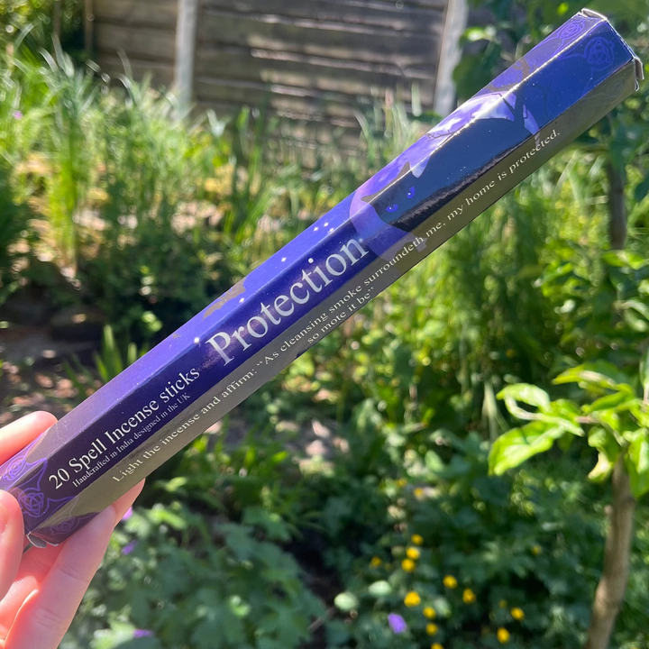 A hand holding a single pack of Elements Protection Spell incense sticks outdoors, showing the deep navy blue and black mystical packaging featuring artwork by Lisa Parker against a lush background of garden foliage to highlight its role in sacred shielding rituals.