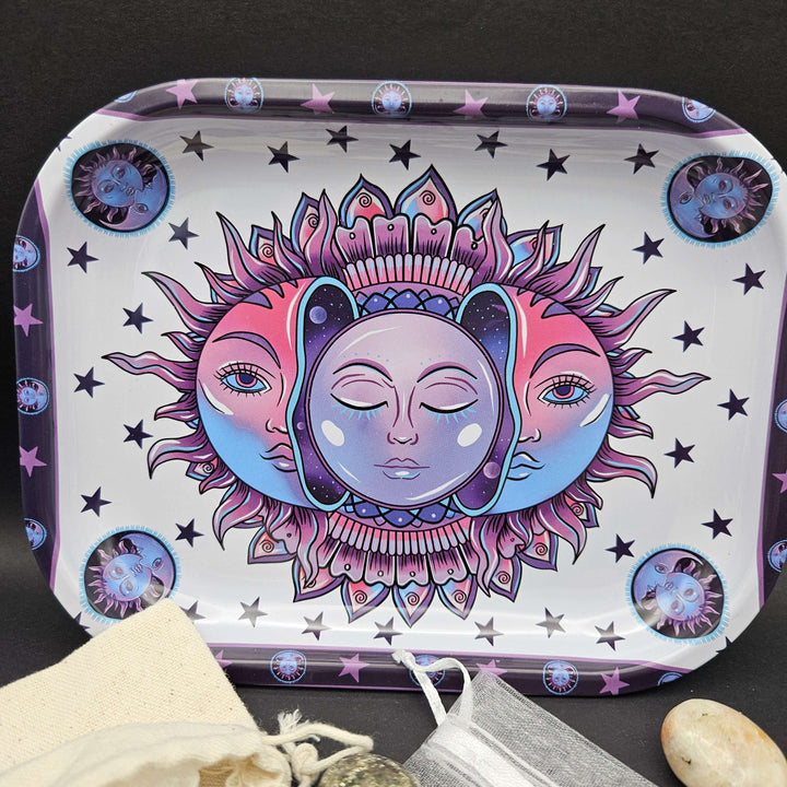 A rectangular metal tin tray with rounded corners decorated with a cosmic sun and moon illustration in pink, blue, and violet. The tray serves as a striking decorative focal point for holding small tumbled stones and ritual accessories. 