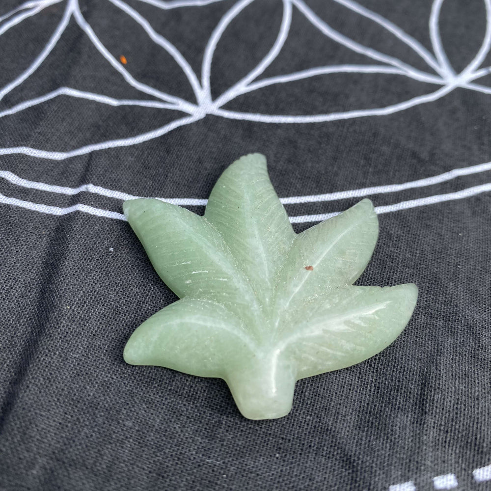 A light green aventurine leaf-shaped pocket stone carving resting centered on a black sacred geometry altar mat printed with white line details.
