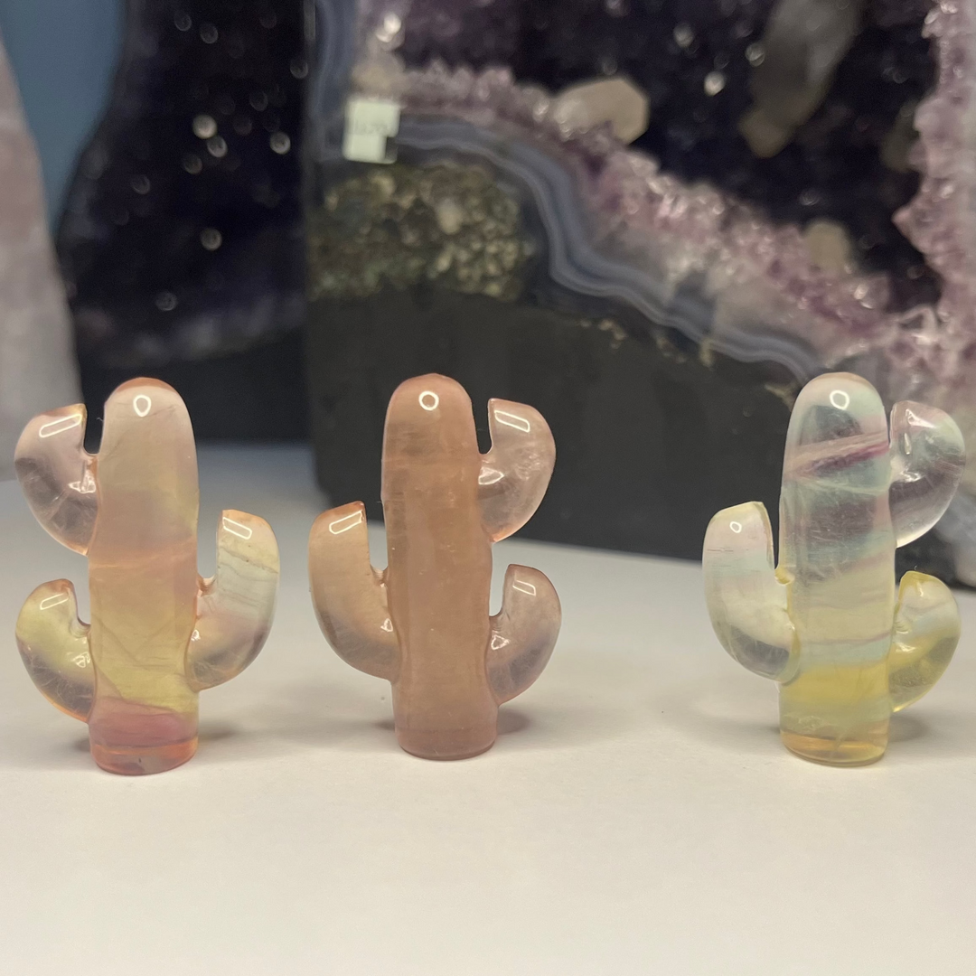 Three mini polished rainbow fluorite cactus crystal carvings standing lined up on a white surface indoors, showcasing translucent naturally occurring colour bands of purple, soft pink, and pale yellow against a large amethyst geode background for Third Eye Chakra alignment.
