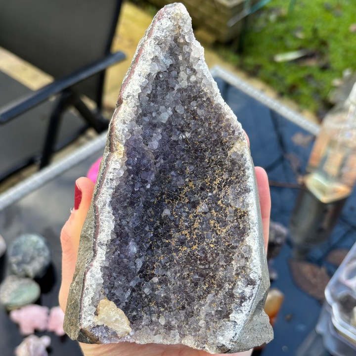 A close-up view of a large, upright raw Amethyst freeform crystal geode chunk held outdoors. The stone showcases a deep cavern face packed with tiny dark purple druzy points framed by a thick white quartz border and rough, natural stone edges.