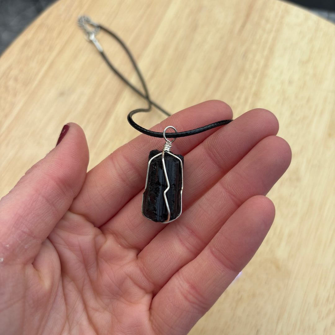 An open hand cradling the raw black tourmaline crystal pendant on its cord over a light wooden table layout, illustrating the compact physical sizing scale of this intuitively chosen security talisman for Root Chakra alignment.

