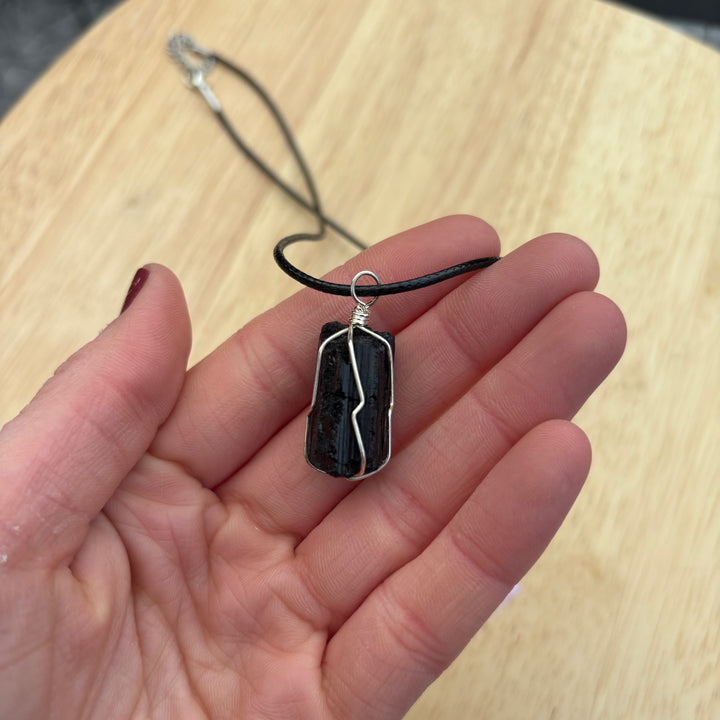 An open hand cradling the raw black tourmaline crystal pendant on its cord over a light wooden table layout, illustrating the compact physical sizing scale of this intuitively chosen security talisman for Root Chakra alignment.
