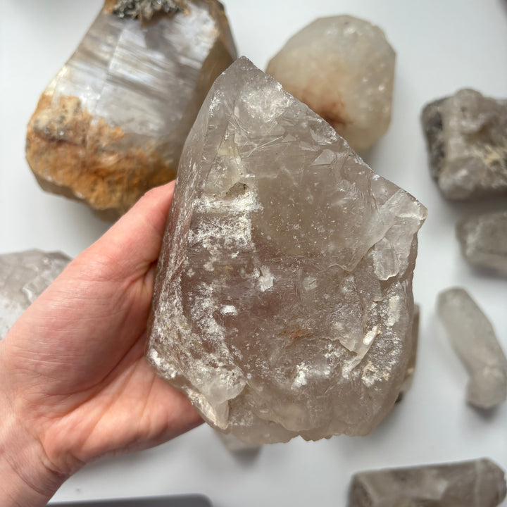 A hand supporting a massive 890g raw clear quartz statement crystal specimen over a white surface scattered with smaller rough minerals, highlighting its unpolished icy frost texture and grand geometric planes for Crown Chakra alignment.