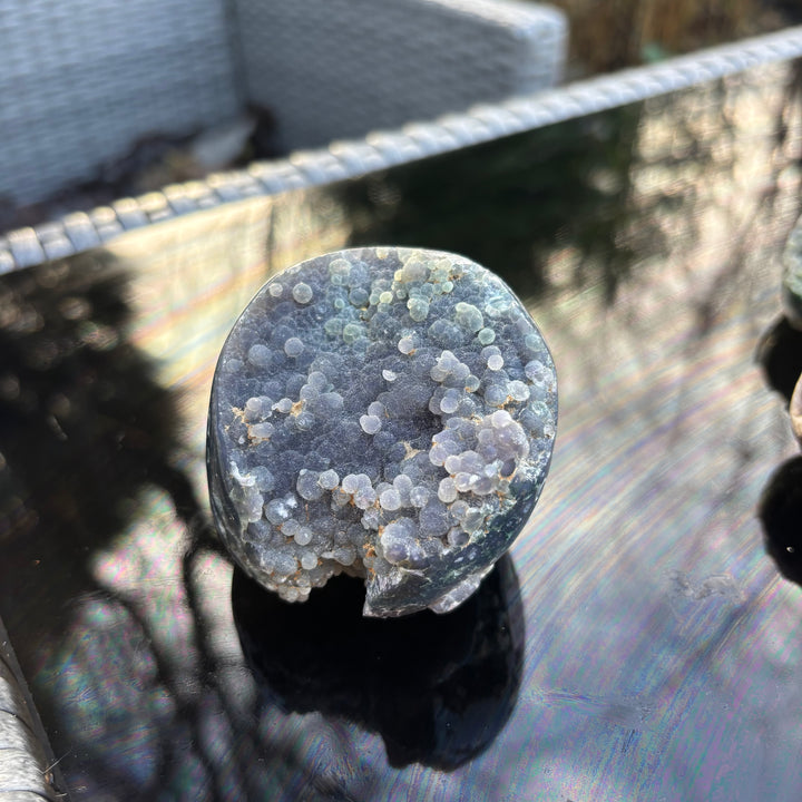 A round purple Grape Agate crystal freeform specimen sitting upright on a black glossy table, catching natural outdoor light.
