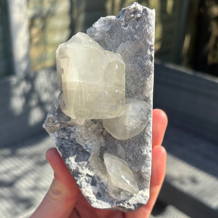 A hand supporting a tall raw grey chalcedony cluster specimen featuring large, semi-translucent clear calcite points growing on a silvery matrix, held outdoors under natural light to showcase its unique 9cm height for Throat and Crown Chakra alignment.
