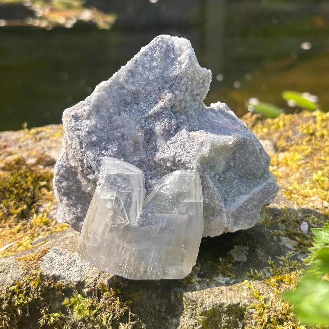 A raw grey chalcedony and clear calcite crystal cluster sitting on a mossy rock outdoors next to a garden pond, showing its shimmering, unpolished textures catching bright natural sunlight for Throat and Crown Chakra balancing.