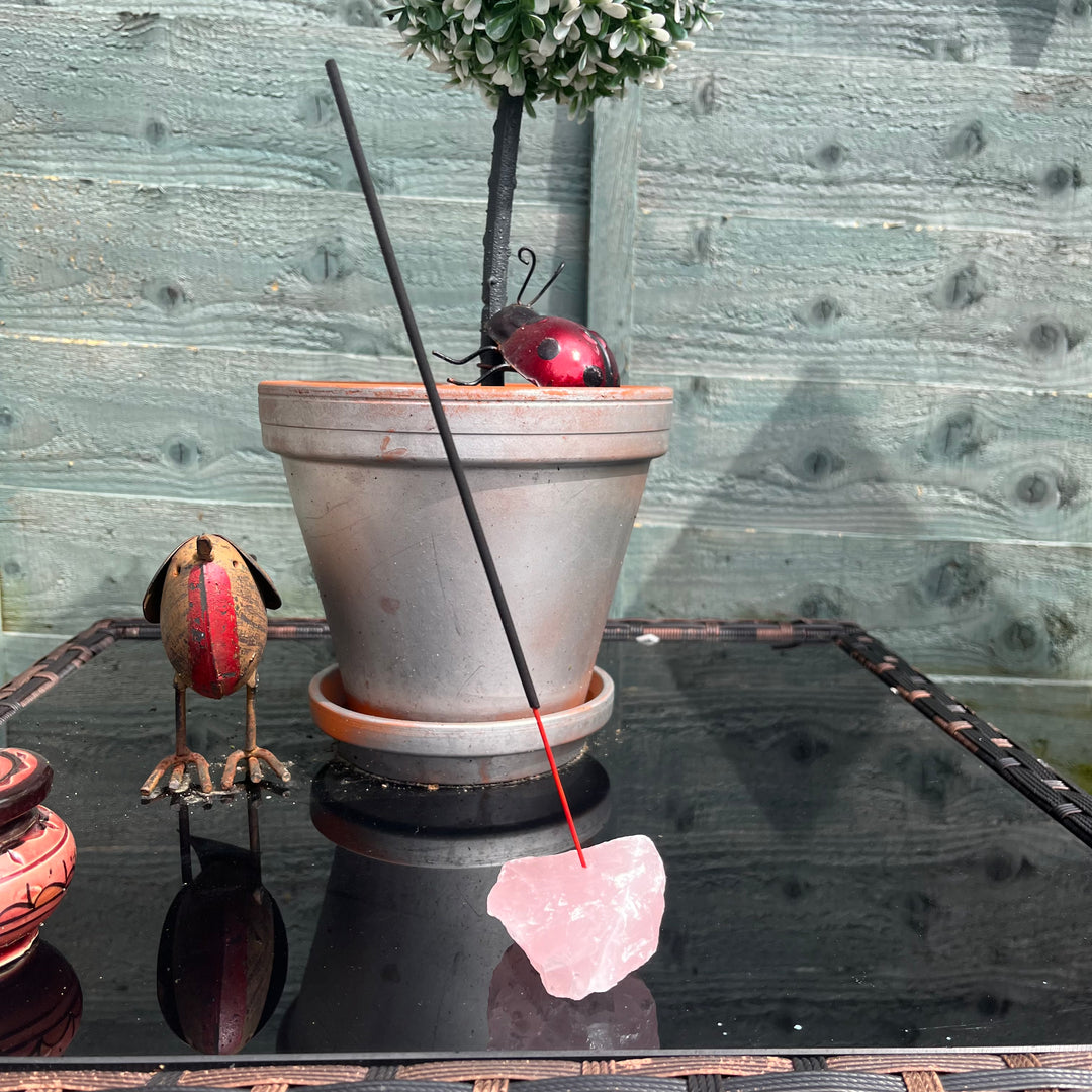 A close-up view of the raw Rose Quartz crystal incense stick holder holding a burning incense stick on a dark glass table. The unpolished blush-pink gemstone chunk displays beautiful earthy layers, natural fractures, and a sharp reflection below.