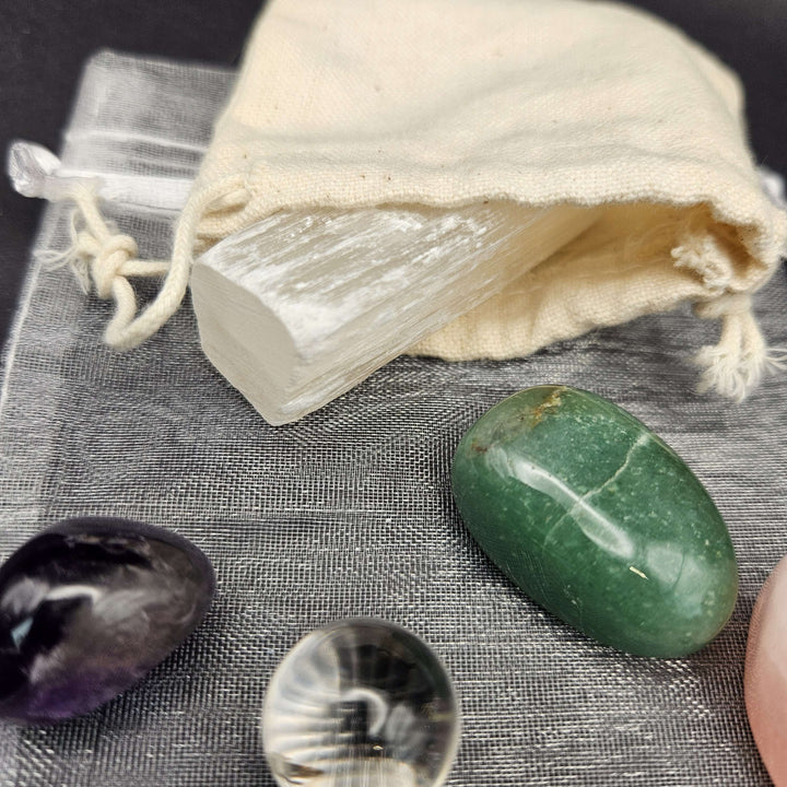 A detailed macro view showcasing a natural white raw Selenite clearing stick tucked neatly inside a cream cotton drawstring pouch. Polished green aventurine and purple amethyst tumbled stones flank the bag on a soft grey surface. 