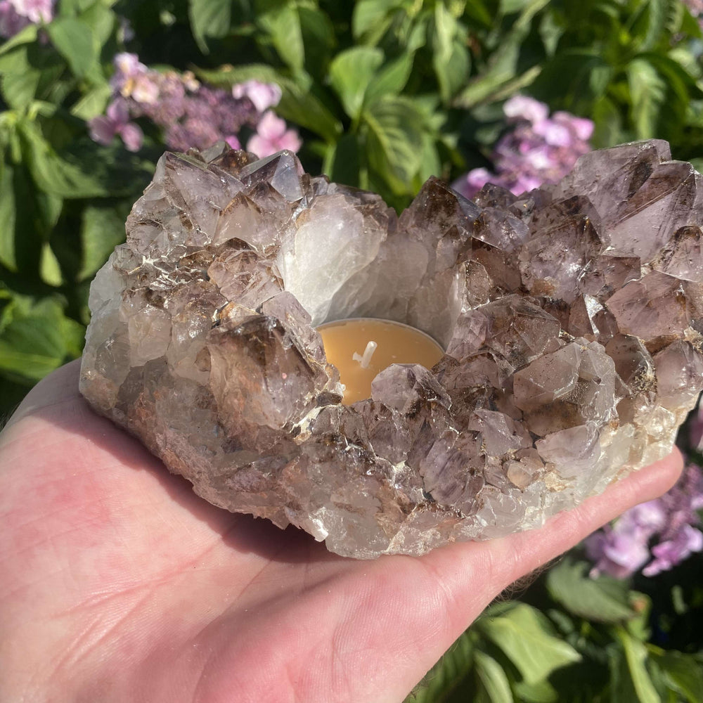 A straight-on handheld view of the raw Spirit Quartz candle holder cluster photographed outdoors. The natural light accentuates the pale smoky-purple quartz points surrounding the central tea light cup, showcasing its unique geological formation.
