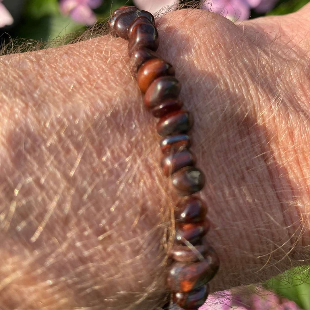 A Red Tiger Eye crystal chip bracelet worn on a wrist outdoors, showcasing the smooth, polished gemstone chips with their rich bands of wine red and deep mahogany tones against garden foliage.