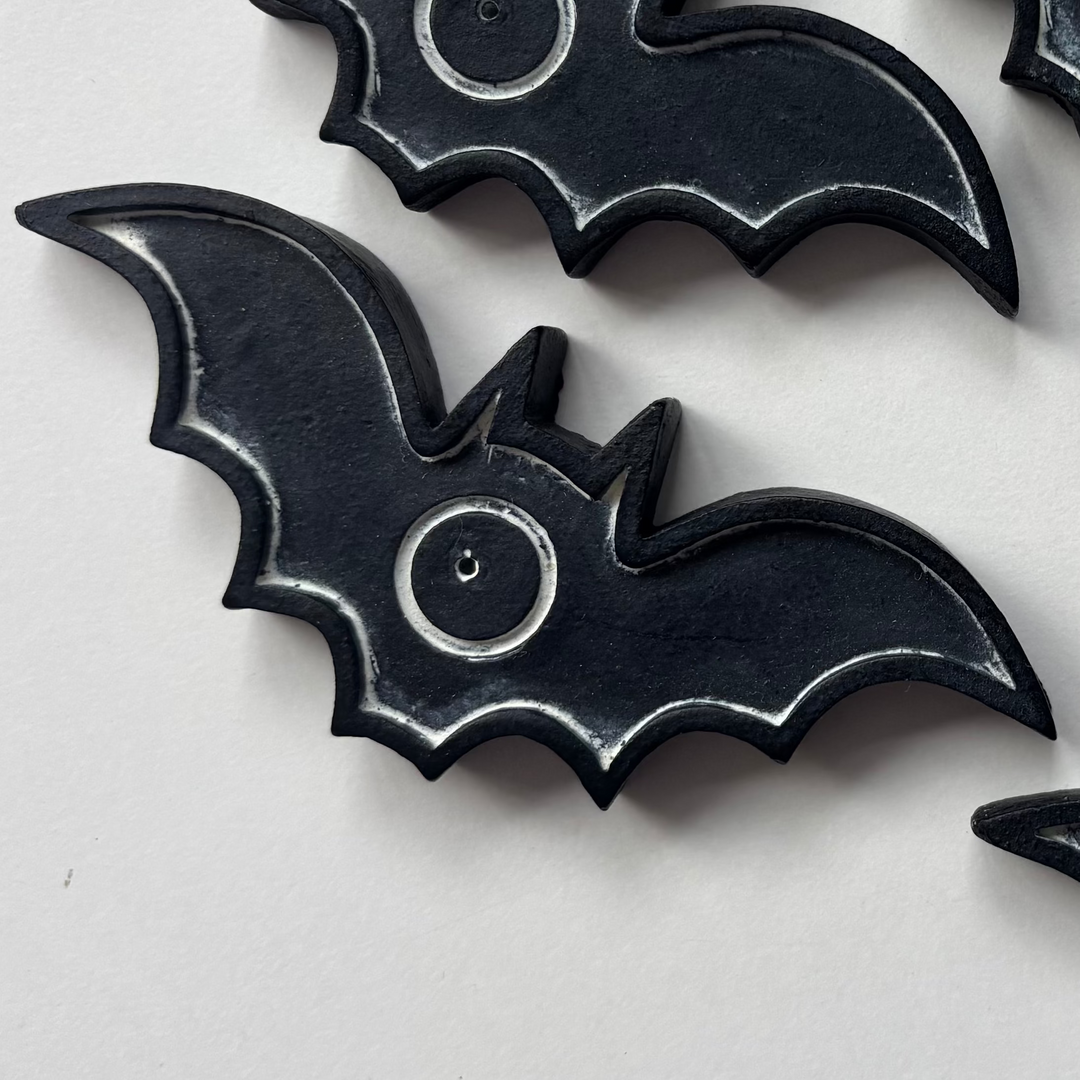 A crisp close-up shot showcasing a cast resin bat-shaped incense burner on a clean white surface. The gothic holder features a smooth matte black finish with carved wing rib accents and a central circular indentation with a pre-drilled incense stick placement hole, styled alongside overlapping duplicate pieces.