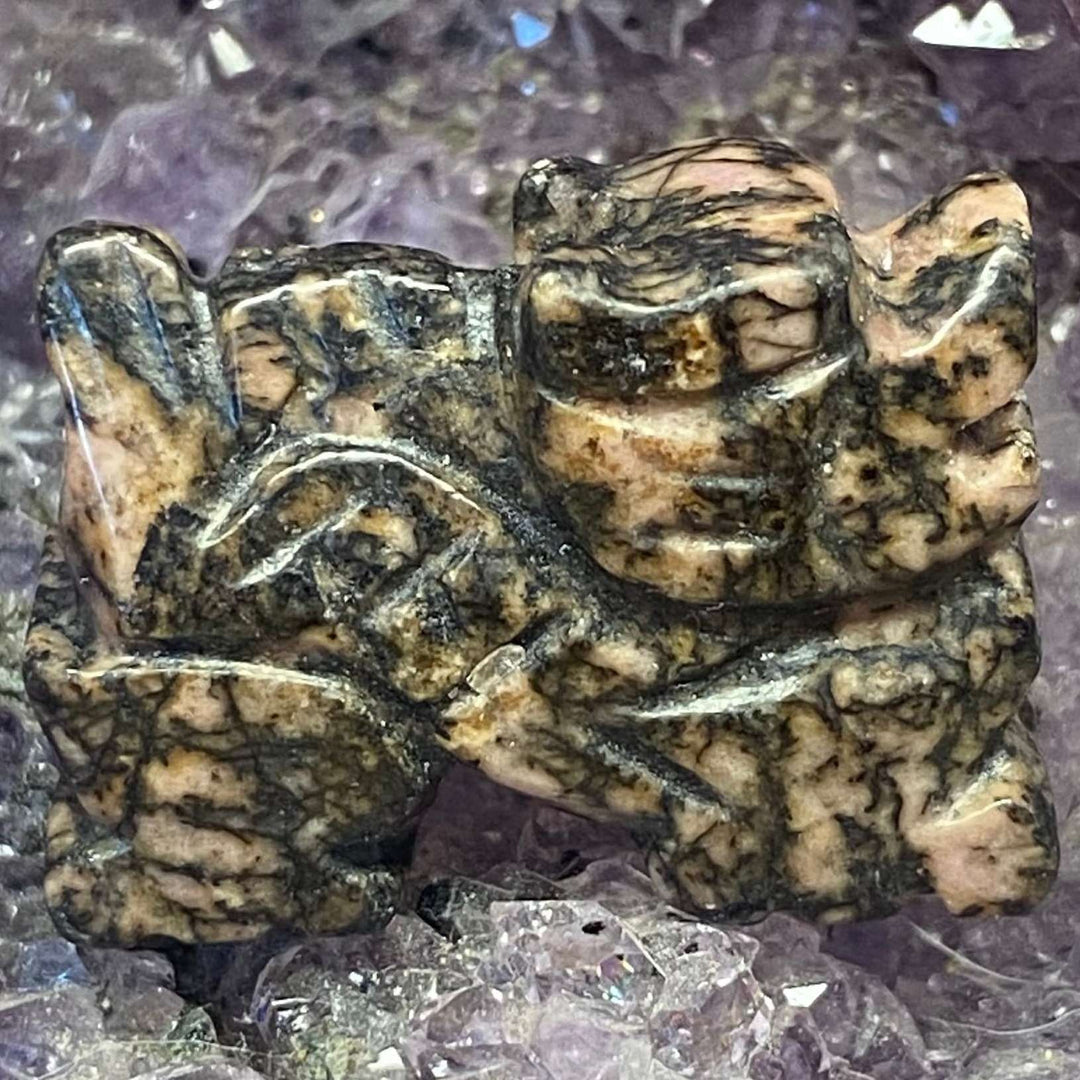 A close-up view of a hand-carved rhodonite dragon crystal, showing off a mottled tan, black, and beige marbled pattern with subtle blue-green veining. The pocket-sized dragon guardian rests on a bed of raw purple amethyst clusters, symbolising soft courage and heart-led power.