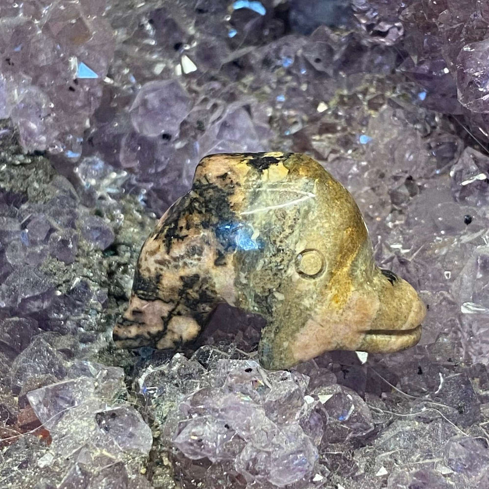 A slightly angled side view of a polished rhodonite gemstone dolphin figurine highlighting its smooth contours and natural stone patterns on a bed of purple crystals.
