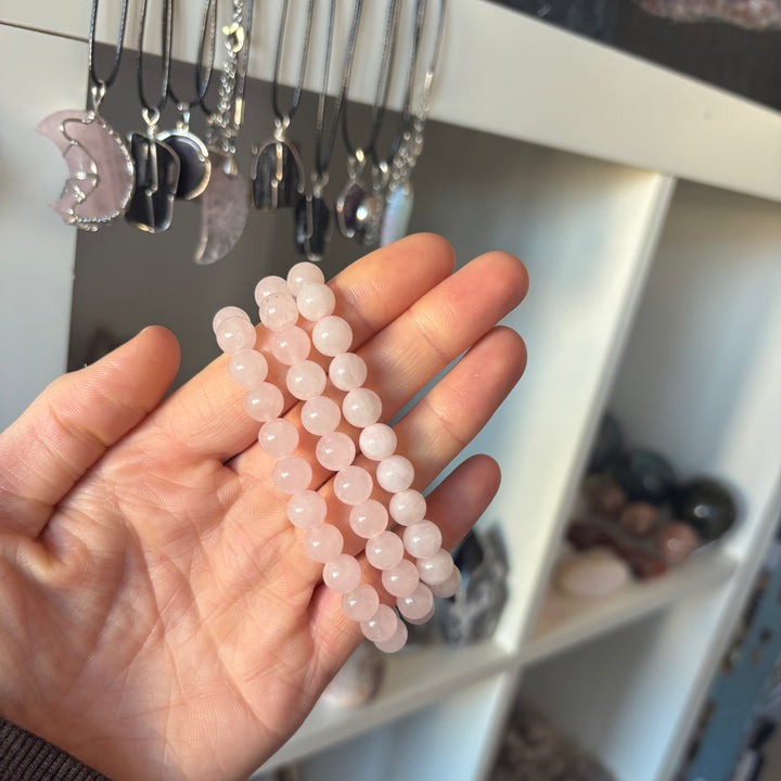 A close-up view of three rounded pink quartz bracelets gathered in a palm inside the boutique. In the background, delicate crystal necklaces softly blur on retail hangers, emphasizing a cohesive, high-quality jewellery presentation.