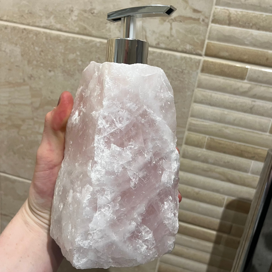 A close-up shot of a substantial raw Rose Quartz crystal soap dispenser held up to view in a person's hand. The heavy block displays a rugged, unpolished chunks stone texture with soft icy-pink colour palettes and internal white quartz crystalline streaks, topped off with a metallic silver pump head against a beige-tiled background.