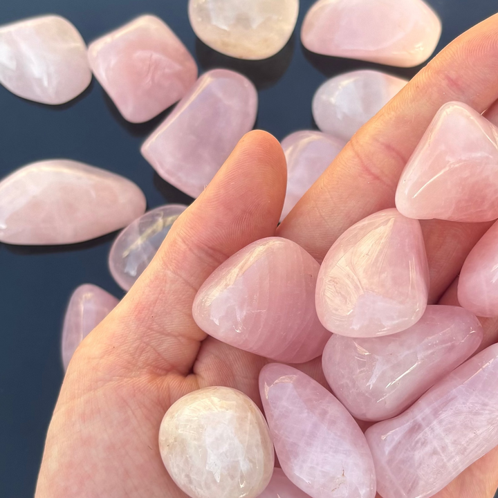 A hand holding a grouping of smooth rose quartz crystal tumbles against a scattered background of polished pink minerals, showing an ideal aesthetic for setting intentions, bedside styling, or a thoughtful wellness gift.