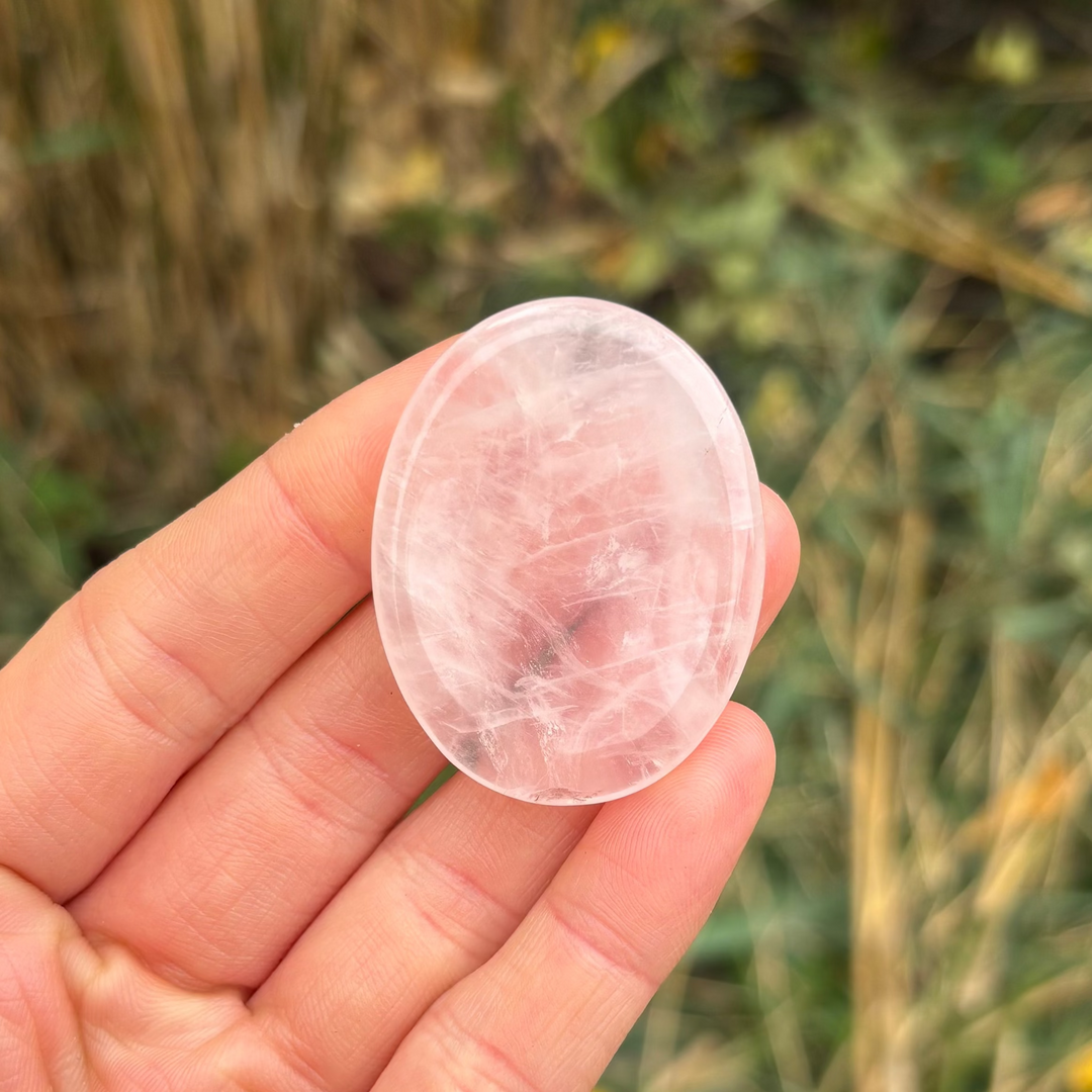 An open hand holding an oval rose quartz crystal worry stone outdoors, showcasing its smooth glassy texture, translucent pale pink clouding, and natural internal veiling against a soft garden background for Heart Chakra alignment.