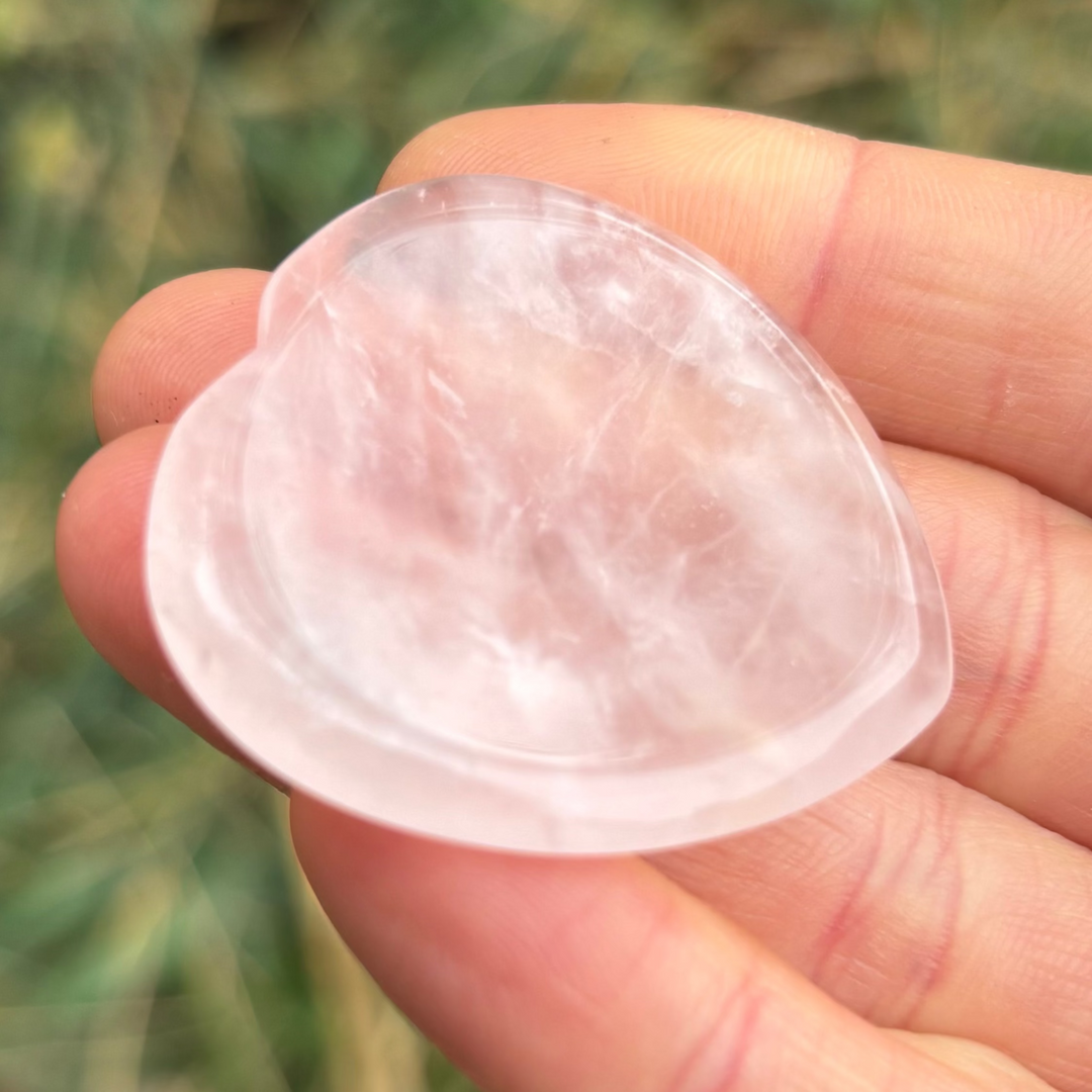 A close-up shot of a smooth, polished pale pink rose quartz heart-shaped worry stone held resting gently on a hand outdoors.