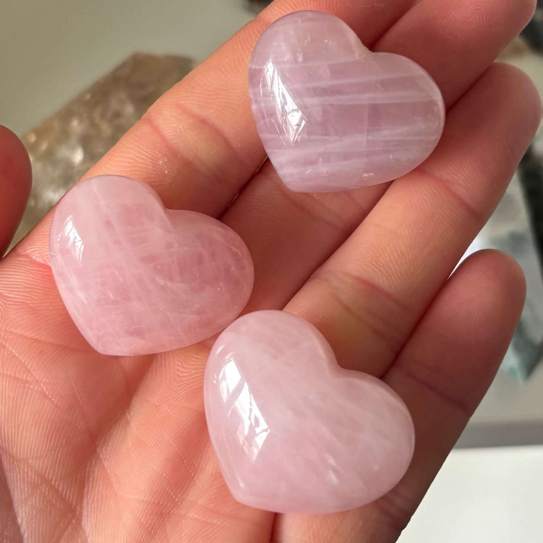 A close-up shot of three smooth, polished pale pink rose quartz crystal hearts held gently in an open palm.