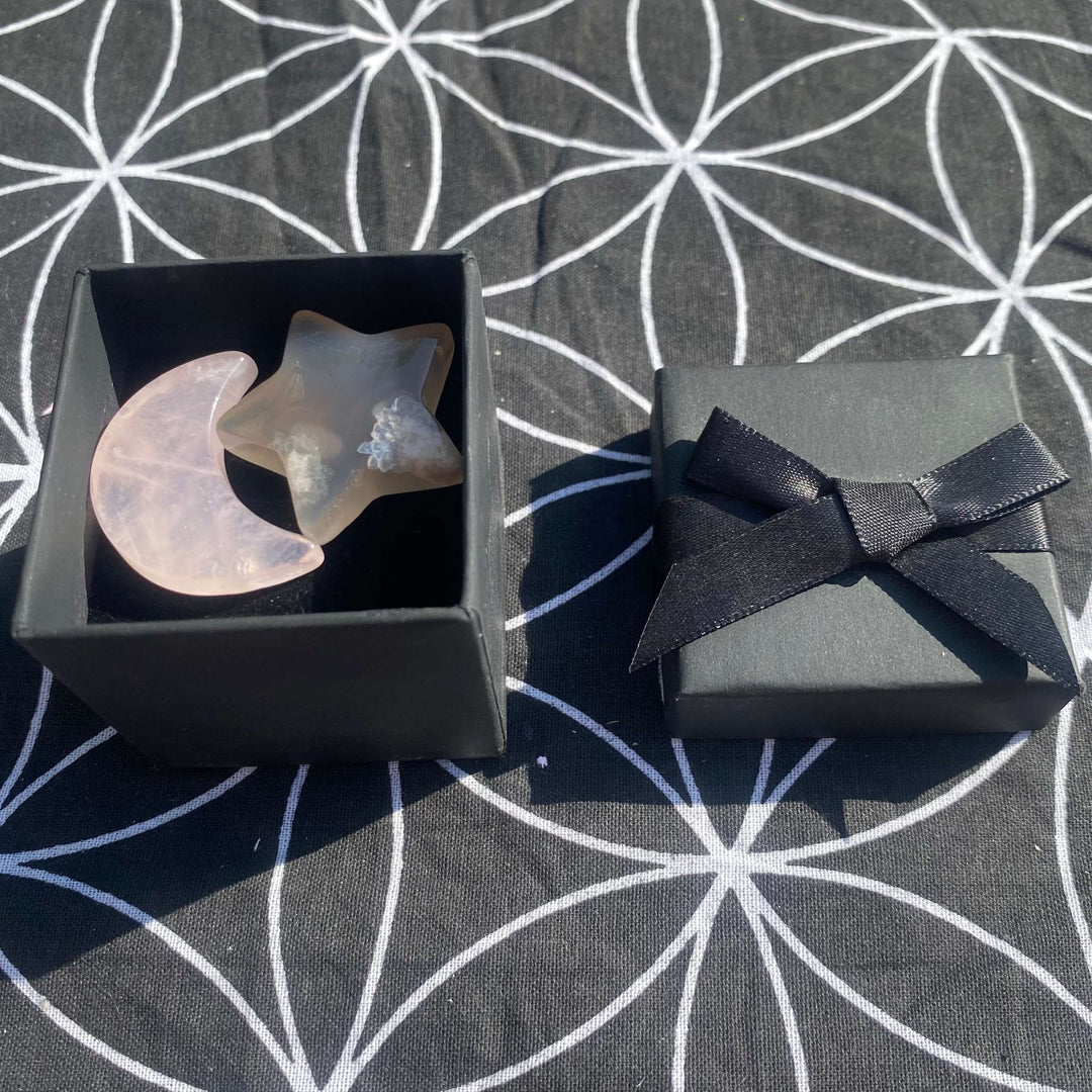 An open textured black square crystal gift box sitting next to its lid on a black sacred geometry mat. Inside the box rests a polished pastel pink rose quartz crescent moon carving alongside a translucent grey agate star pocket stone.