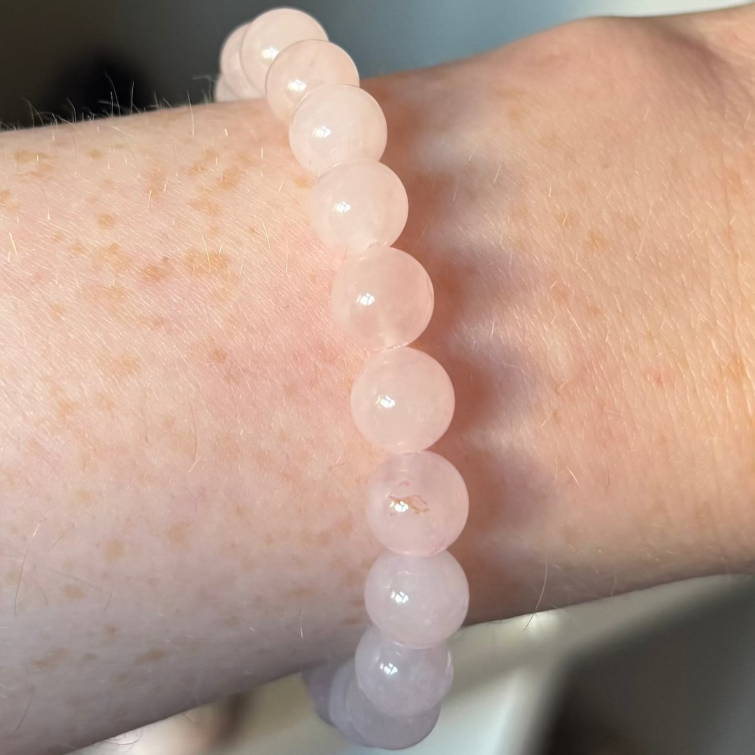 A polished Rose Quartz round beaded bracelet worn on a wrist, showcasing its smooth, perfectly circular pink crystal beads with a gentle translucent quality. The lightweight stretch design contours the skin comfortably against a bright indoor background.