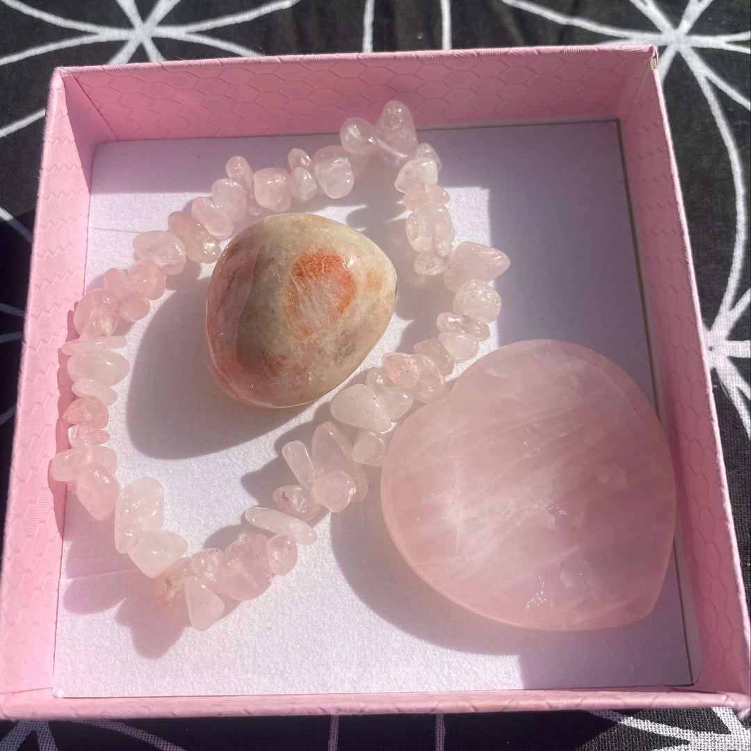 A top-down view looking into an open, square light pink box displaying the premium crystal love gift set. Inside, a delicate blush-pink Rose Quartz chip bracelet encircles a smooth, polished Sunstone tumble stone, resting alongside a large, smooth Heart-shaped Rose Quartz thumb stone on a crisp white insert.
