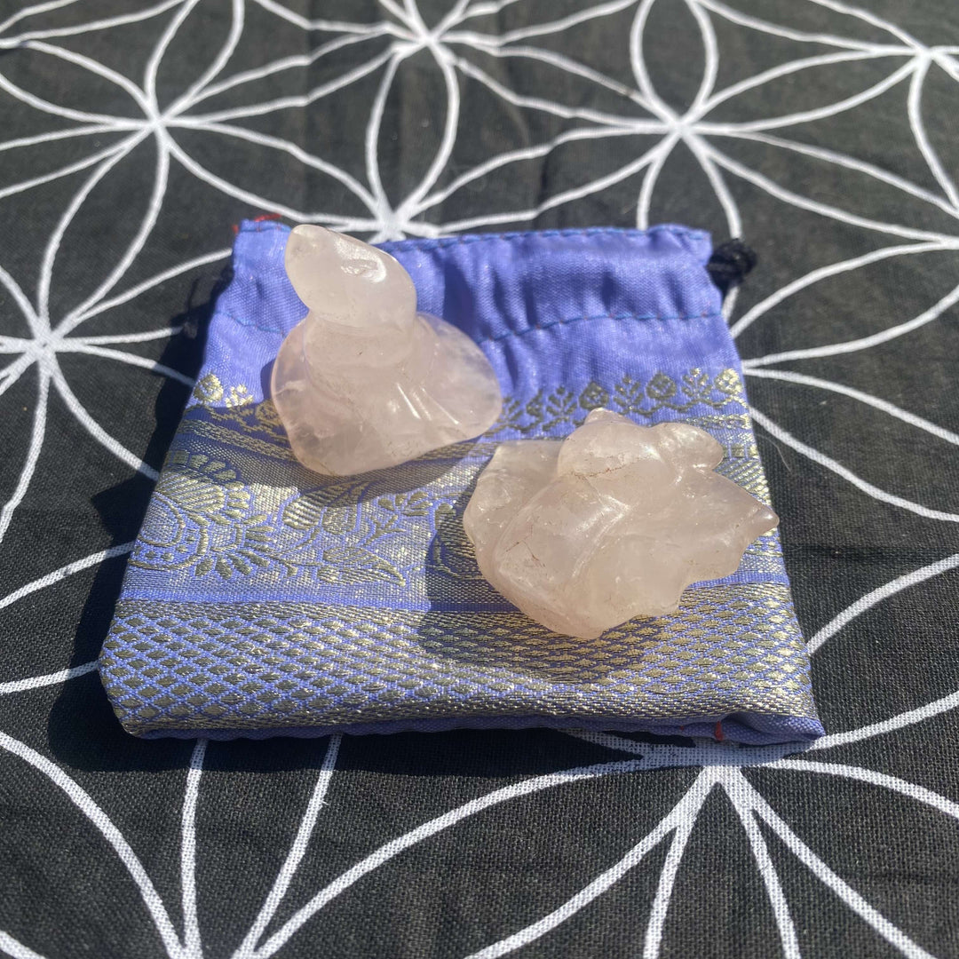 Overhead shot of a hand-carved pale pink Rose Quartz witch's hat and matching bat figurine on a periwinkle blue and silver jacquard drawstring pouch